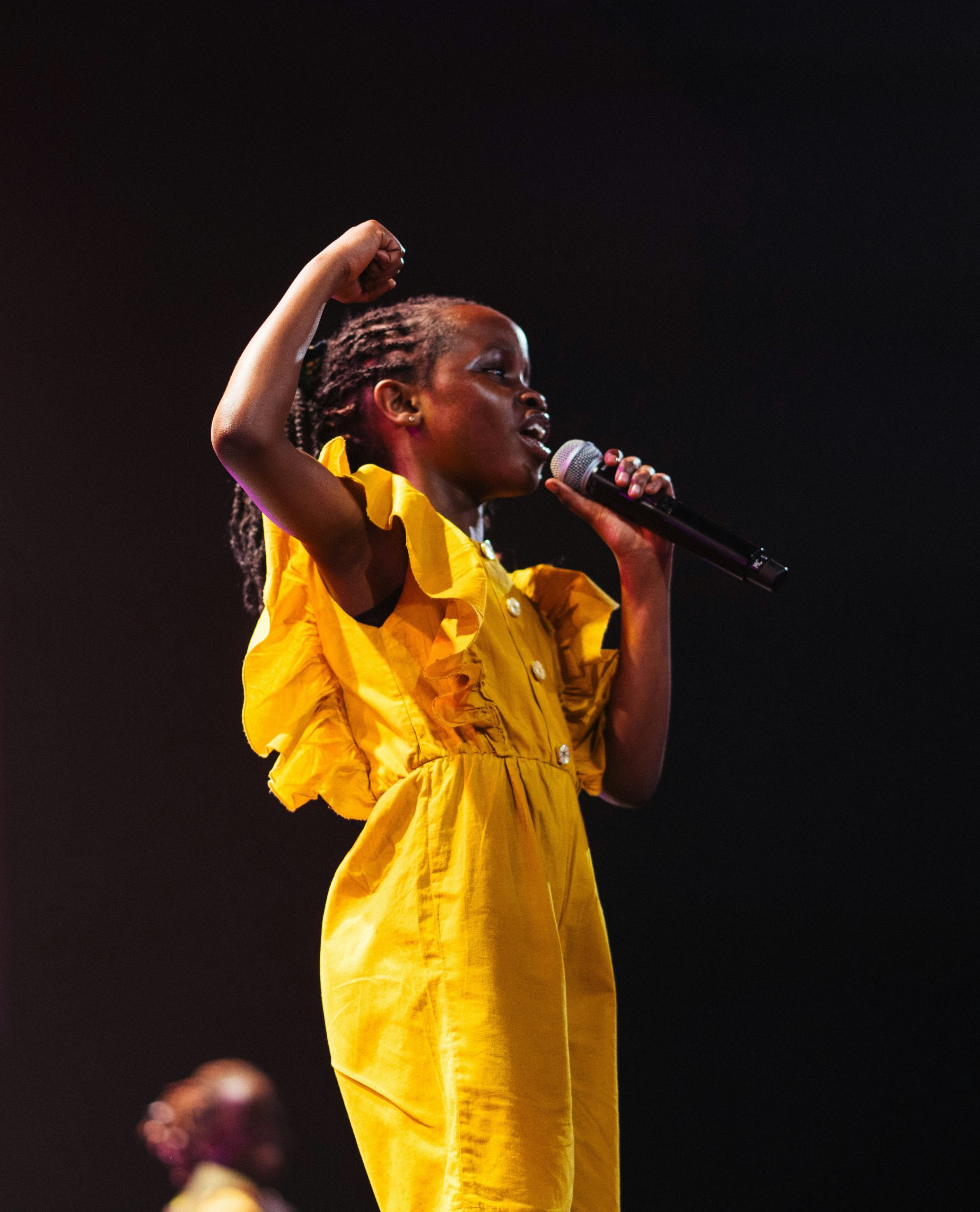 A child in a yellow dress sings into a microphone with her fist raised on stage